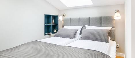 Un dormitorio con tragaluz, cama con ropa de cama gris, mesita de noche y estantería azul.