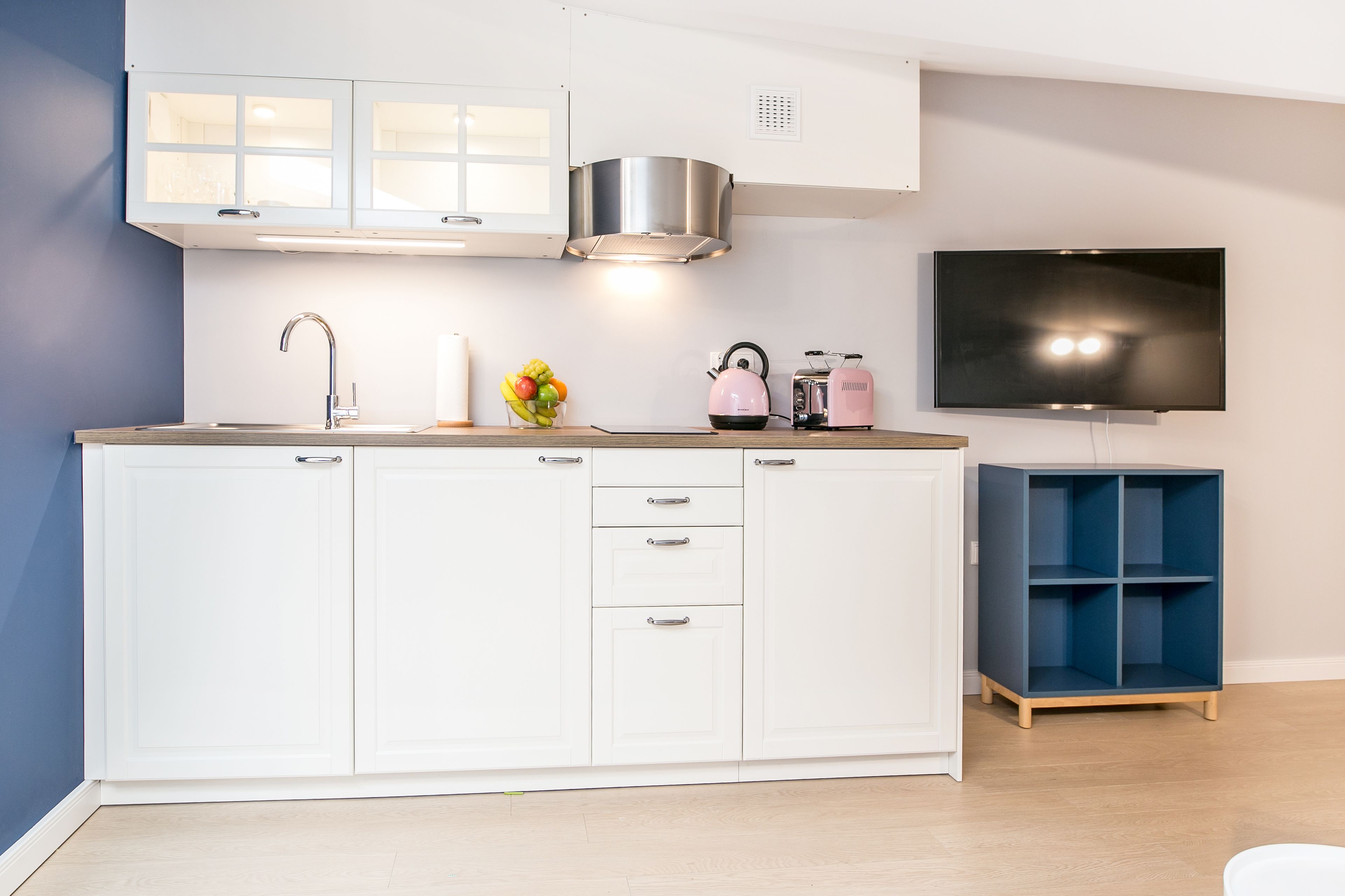 A modern kitchen with white cabinets, a stainless steel range hood, a built-in TV, and a blue shelving unit.