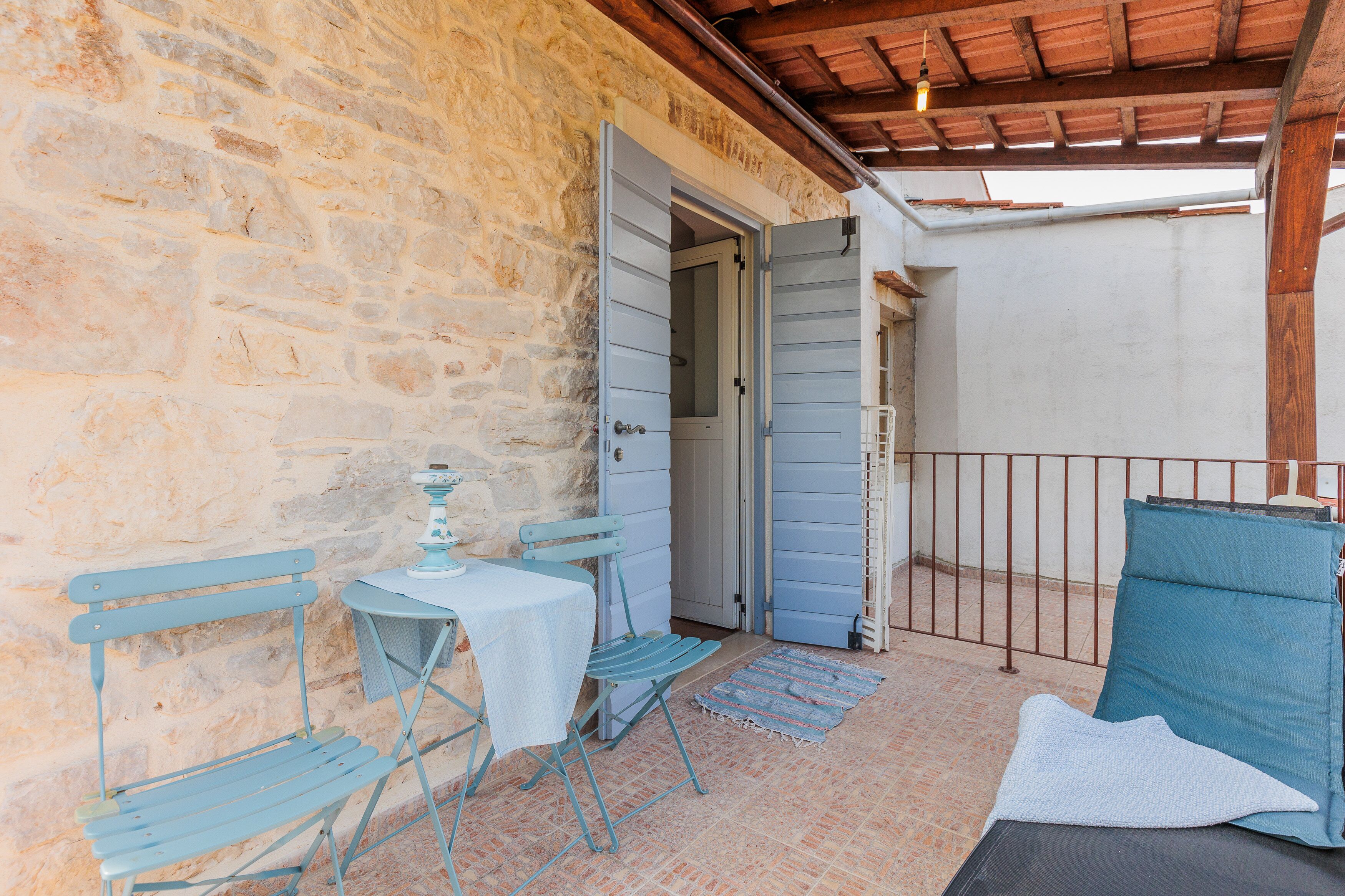 Un espace extérieur entouré de murs en pierre, avec une table et deux chaises, une porte et une rambarde en métal.