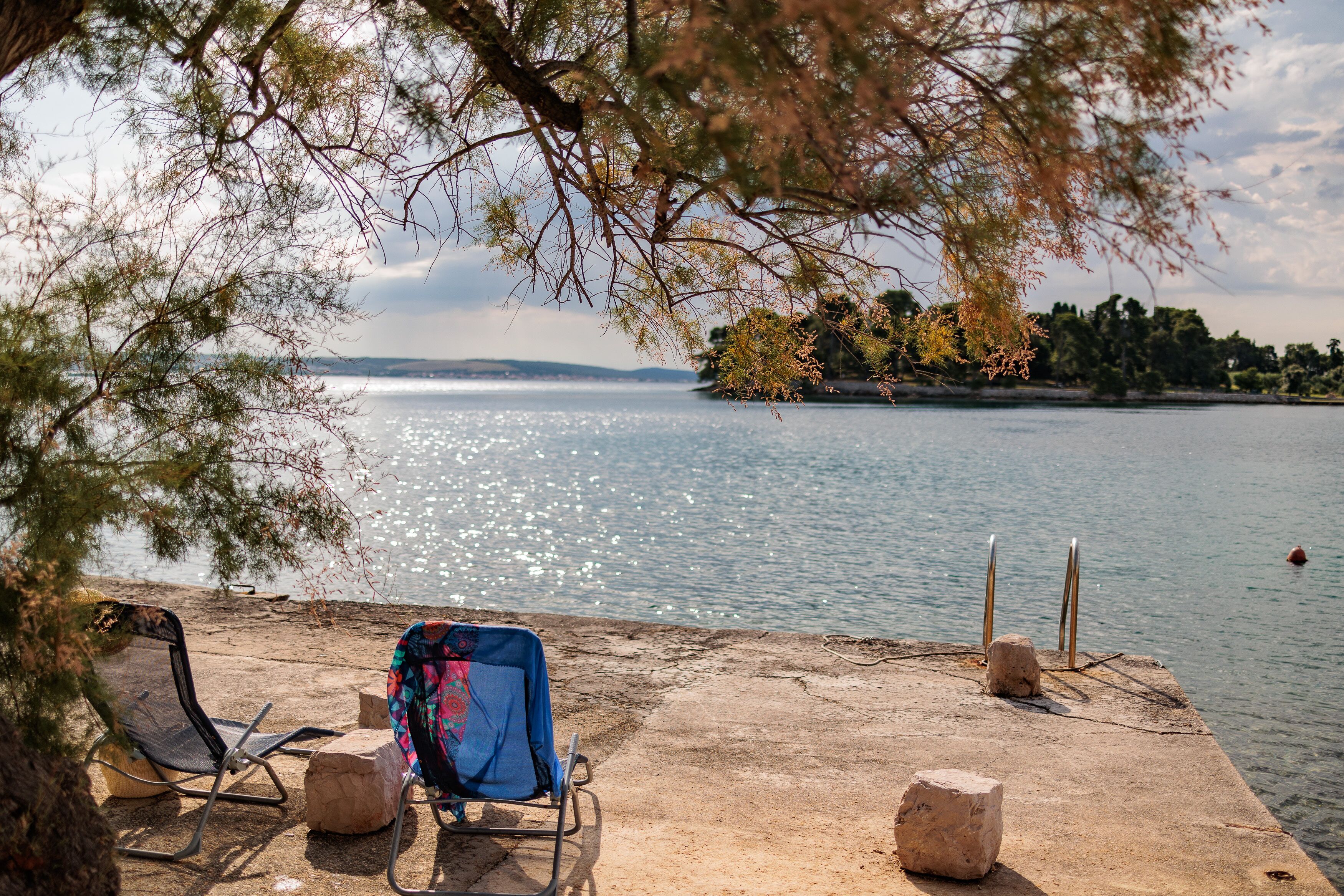 A lakeside view with a concrete dock, two metal chairs, and a covered chair with a colorful blanket.