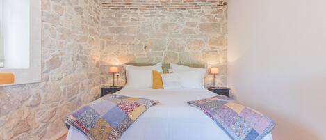 Un dormitorio con pared de piedra, suelo de madera, cama con colcha, dos lámparas de noche y una ventana.