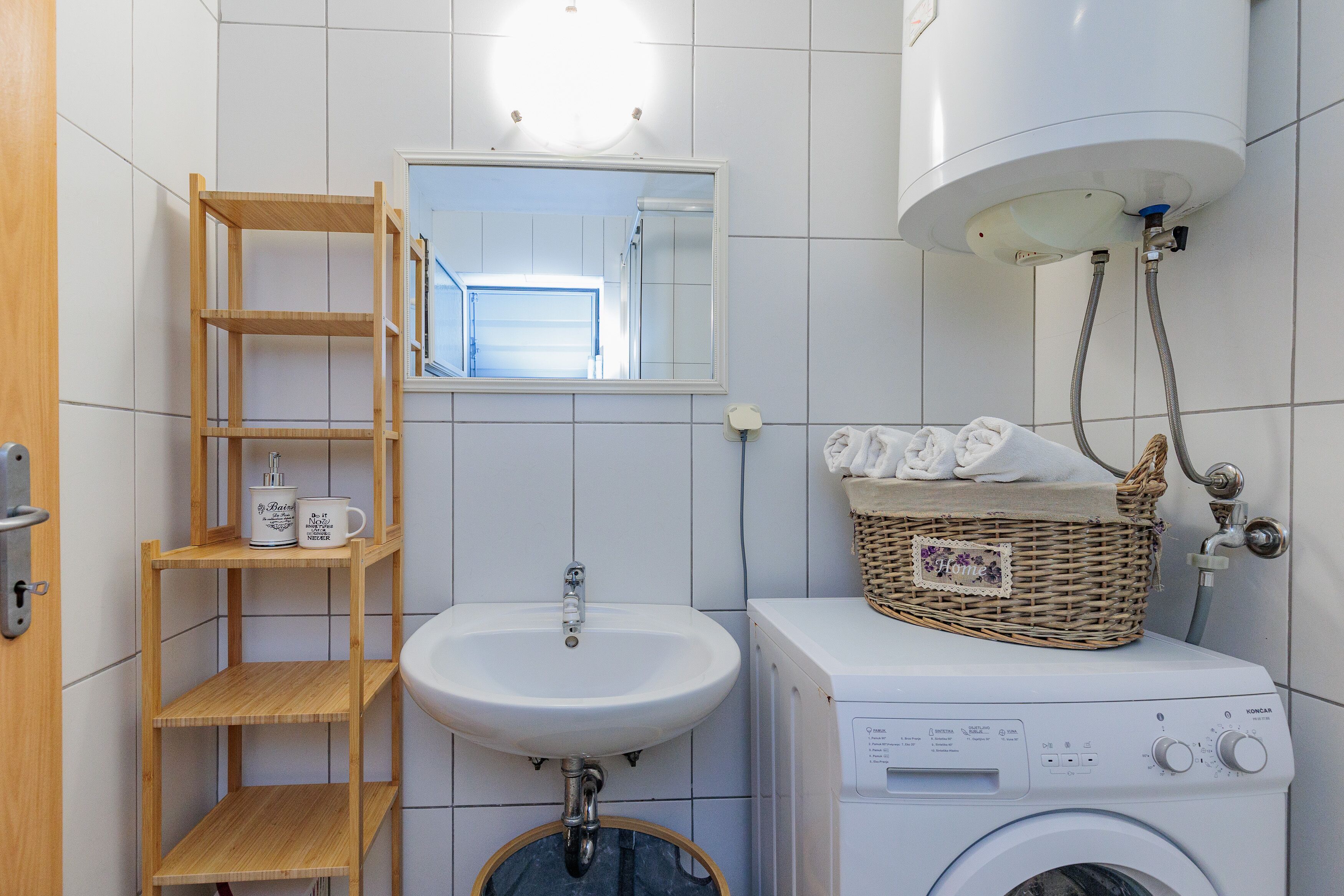 A bathroom with a wooden shelf, a white sink, a mirror, a washing machine, and a basket on the machine.