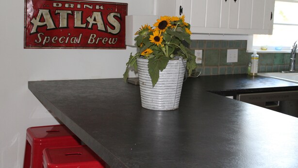 Eine Küchenarbeitsplatte mit einem weißen Eimer voller Sonnenblumen, einem roten Schild mit der Aufschrift 'DRINK ATLAS Special Brew' und weißen Schränken.