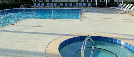 A swimming pool area with lounge chairs and umbrellas, a separate hot tub, and a building in the background.