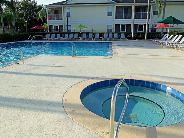 A swimming pool area with lounge chairs and umbrellas, a separate hot tub, and a building in the background.