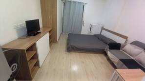 Una stanza compatta con un letto, una scrivania con un computer, una ventola e una lavatrice.