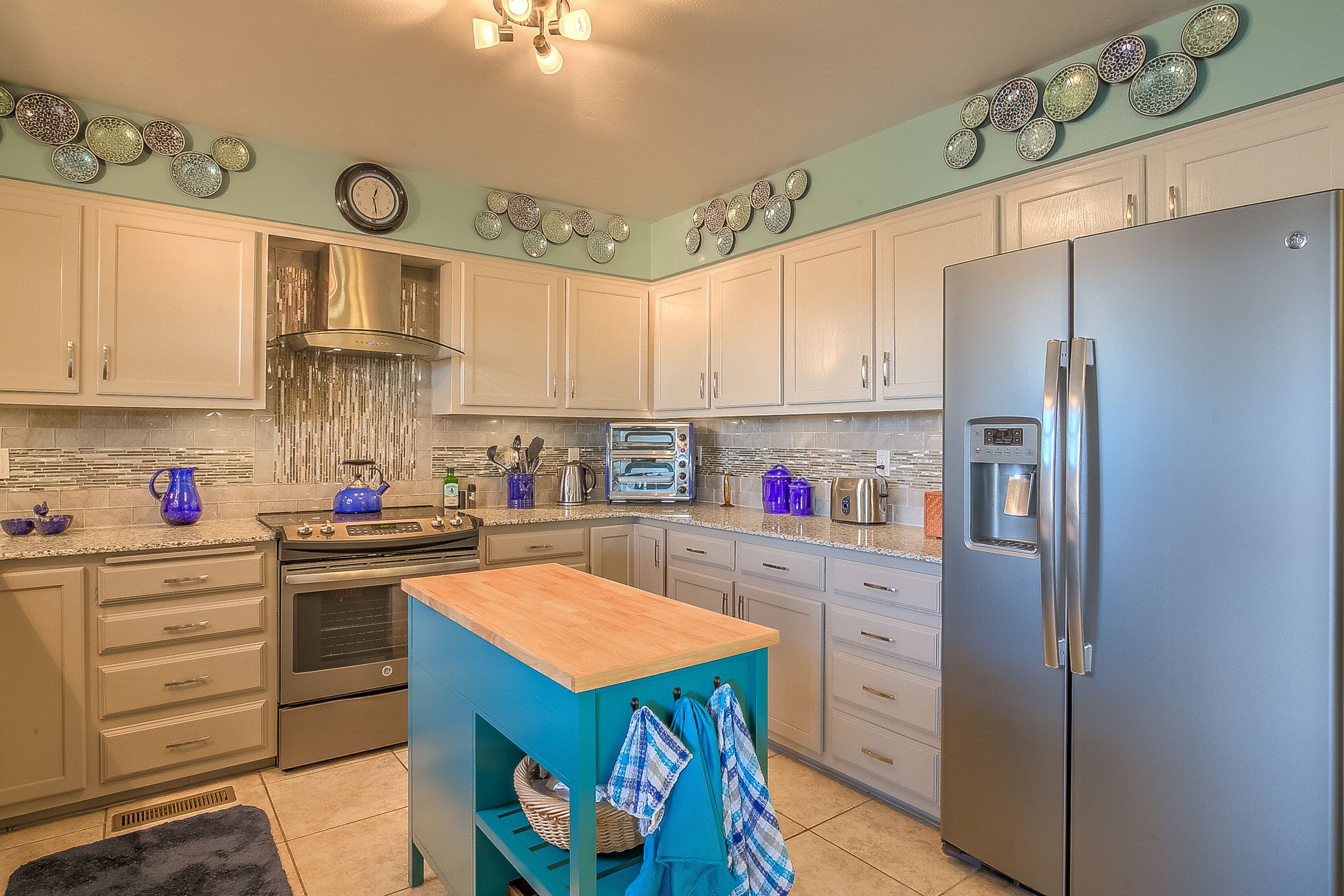 A kitchen with white cabinets, a stainless steel refrigerator, a wooden island, and decorative plates on the wall.