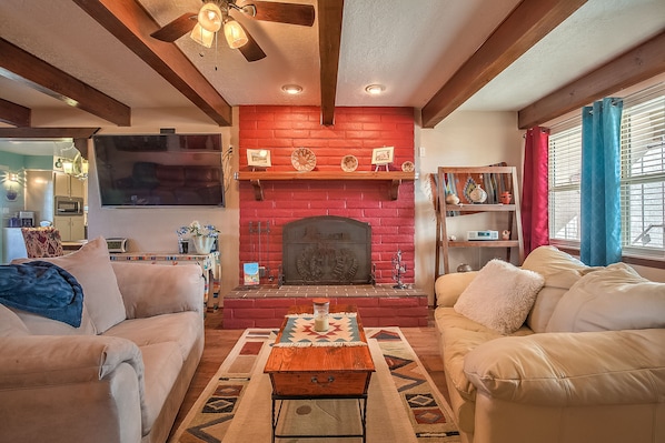 Una sala de estar con chimenea de ladrillo rojo, sofás color beige, una mesa de centro de madera y un ventilador de techo.