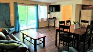A living room with a sofa, a dining table with chairs, a TV mounted on the wall, and a kitchen area with a refrigerator.