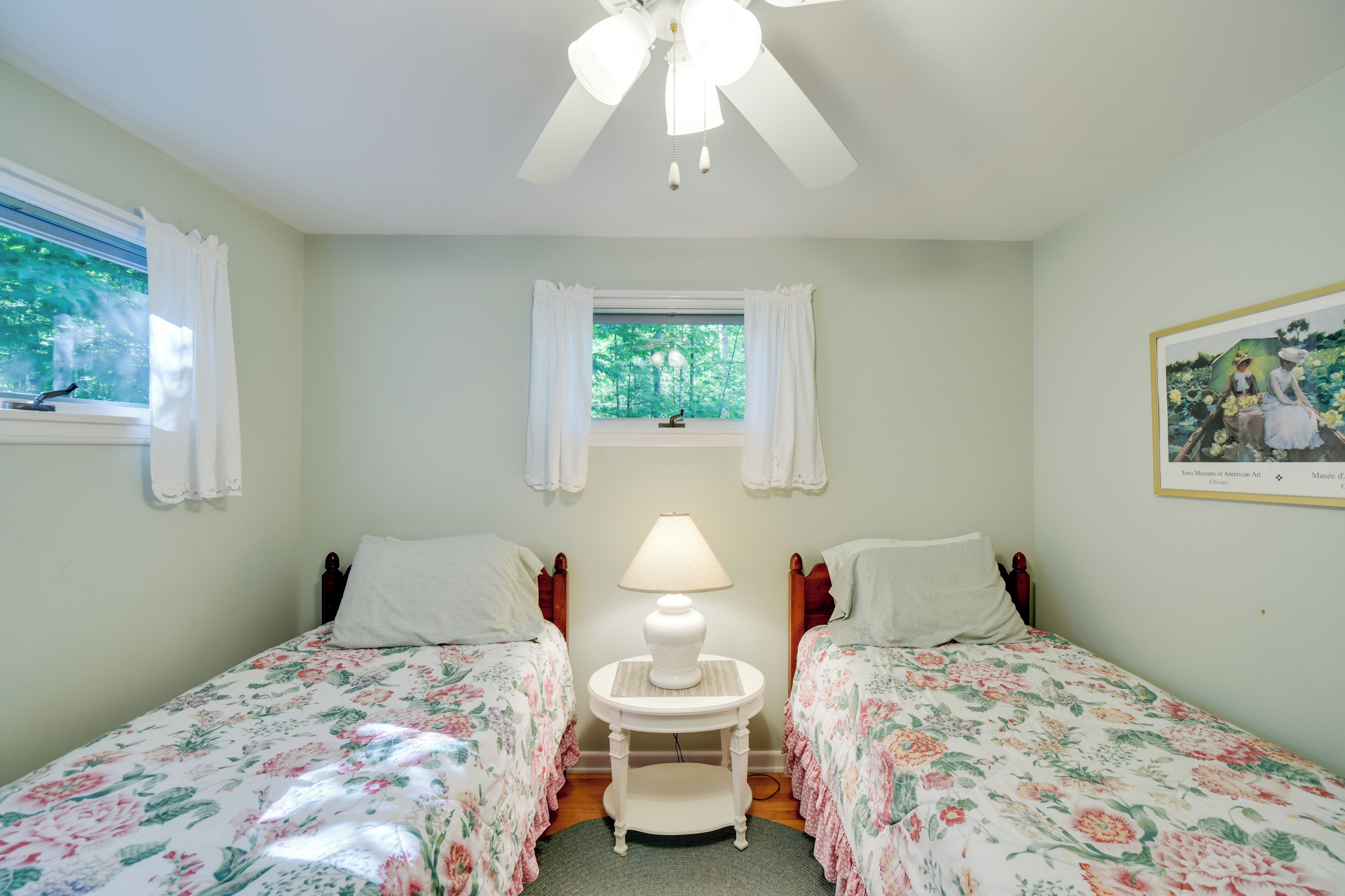 Une chambre avec deux lits simples, un ventilateur de plafond, une table de chevet avec une lampe, une fenêtre avec des rideaux et un tableau encadré au mur.