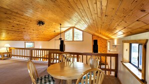Una habitación amplia con techo de madera, una mesa redonda de comedor con sillas, barandilla de madera y una ventana.