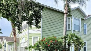 A two-story residential building with a green exterior and white trim, surrounded by well-maintained lawns and a variety of plants.