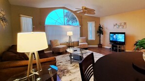 A living room with a sofa, armchair, coffee table, TV, and a dining table with a potted plant.