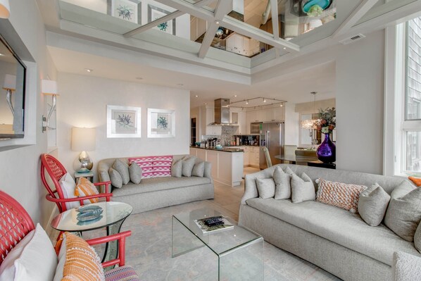 A spacious living area with a high ceiling, a kitchen in the background, and a dining area with a table and chairs.