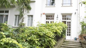 A white building with a staircase leading to a door, surrounded by greenery.