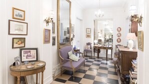 A hallway with a checkered floor, a mirror, a purple armchair, and a table with a vase.