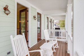 A covered porch with white railings, wooden flooring, and white chairs.
