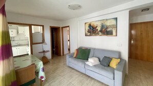 Ein Wohnzimmer mit grauem Sofa, einem Esstisch mit Stuhl, einer Küchenzeile und einer Tür zu einem anderen Raum.