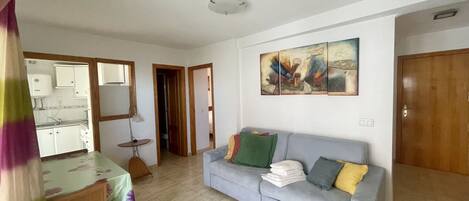 A living room with a grey sofa, a dining table with a chair, a kitchen area, and a door leading to another room.