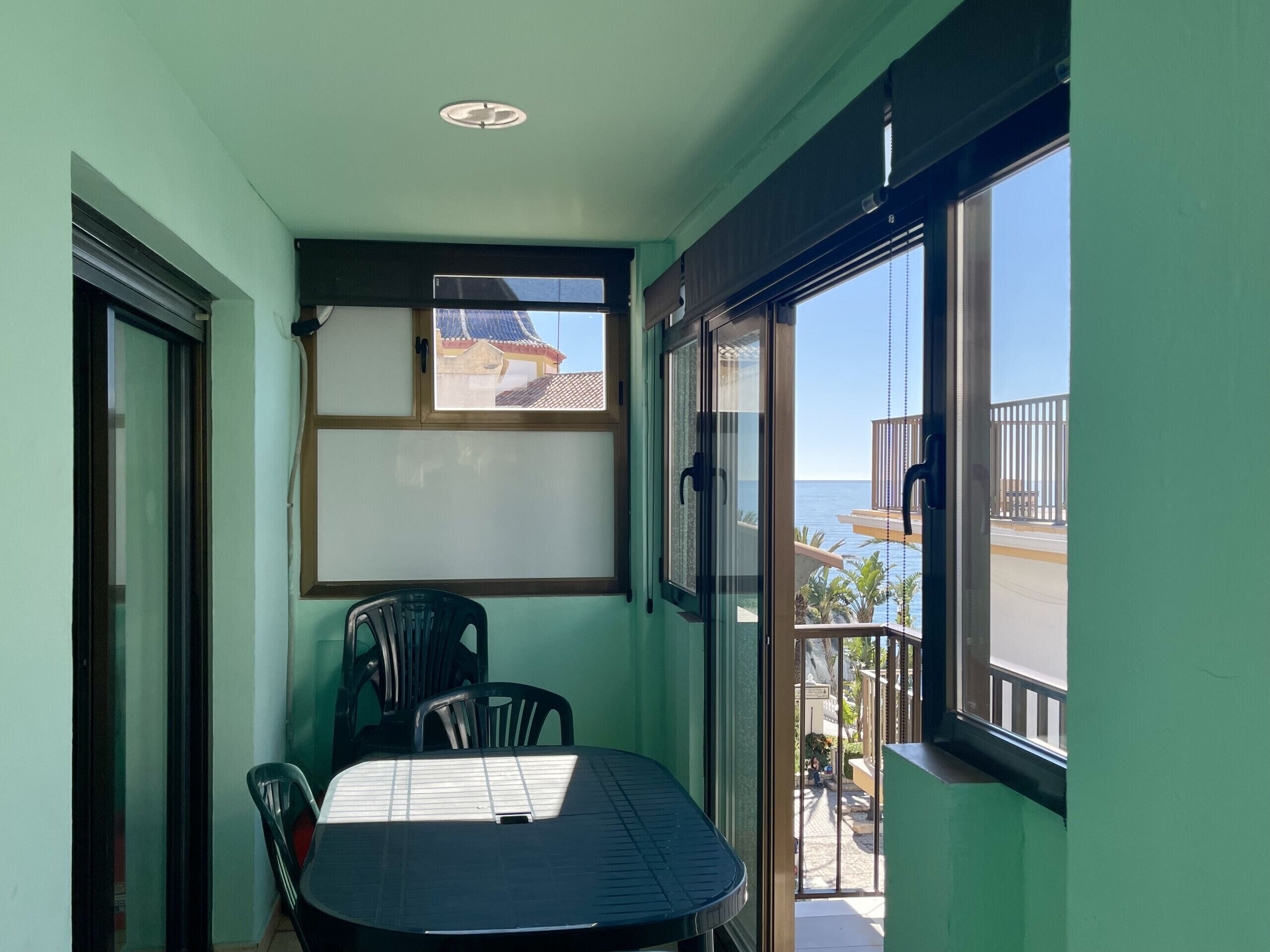 A small balcony with a table and chairs, a view of the sea, and a building with a blue roof in the background.