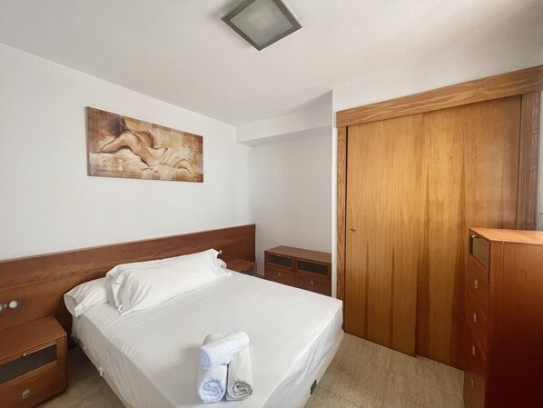 A hotel room with a wooden bed frame, white linens, a painting on the wall, and a wooden wardrobe.