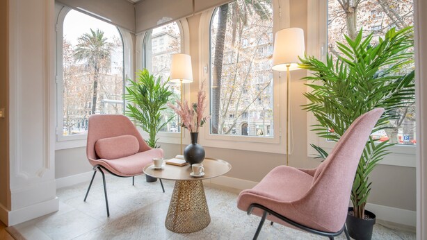 Een moderne kamer met twee roze stoelen, een ronde tafel en grote ramen met uitzicht op bomen en gebouwen.