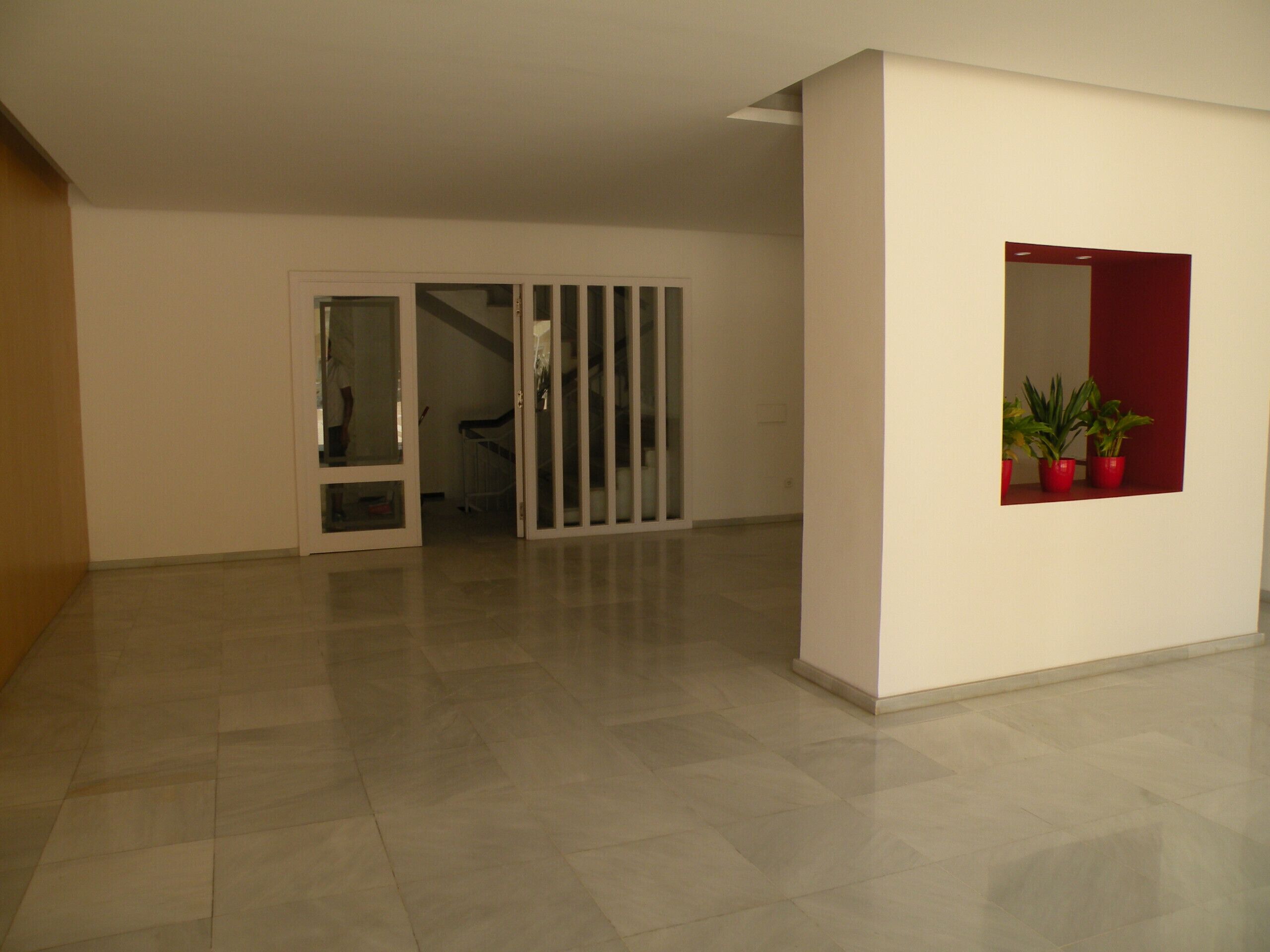 Un pasillo amplio y luminoso con suelo de baldosas, una puerta corredera de vidrio que da a otra habitación y un ventanal con plantas en macetas.