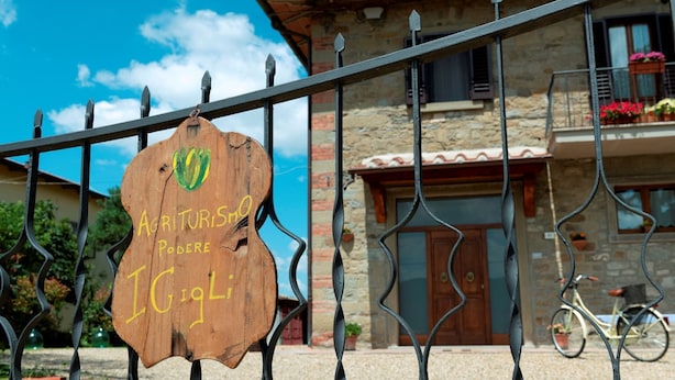 A wooden sign with text, a metal fence, and a building with a stone facade and a bicycle. - Recently restored stone villa with private pool, ideal for couples of friends or families wishing to (Castiglion Fiorentino)