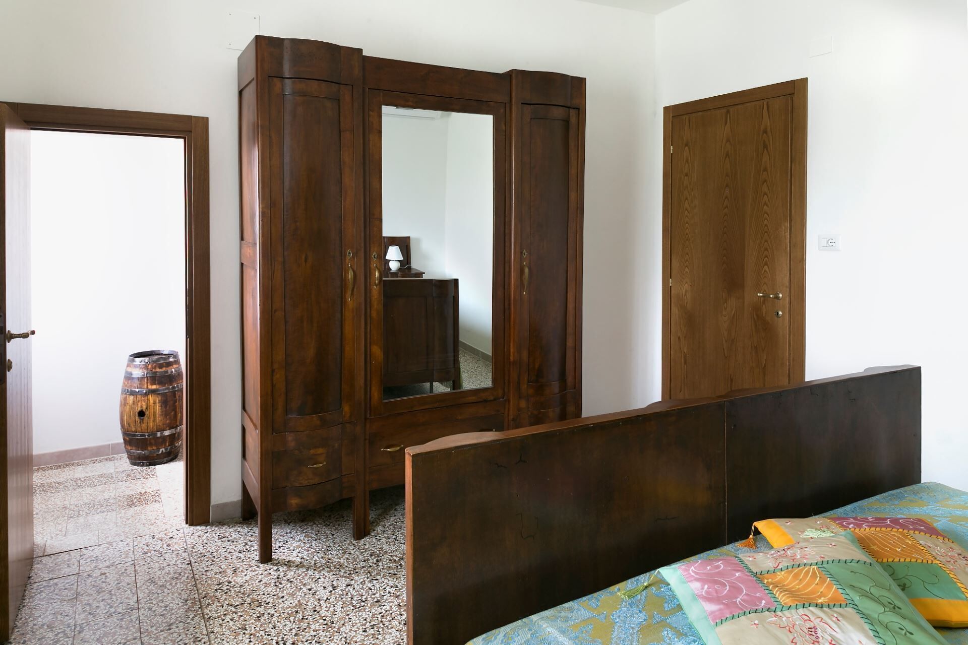 Une chambre avec une armoire en bois, un meuble avec miroir, un lit recouvert d’une courtepointe colorée et un tonneau en bois posé sur le sol.