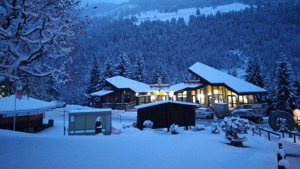 Un chalet enneigé composé de plusieurs bâtiments, dont un bâtiment principal et de plus petits chalets, le tout entouré d’une montagne boisée.
