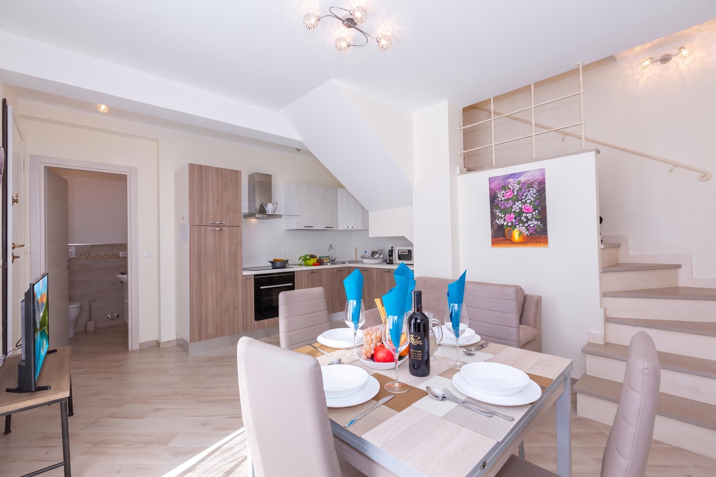 Una cucina moderna con mobili in legno, una zona pranzo con tavolo apparecchiato per quattro persone e una scala che conduce a un piano superiore.
