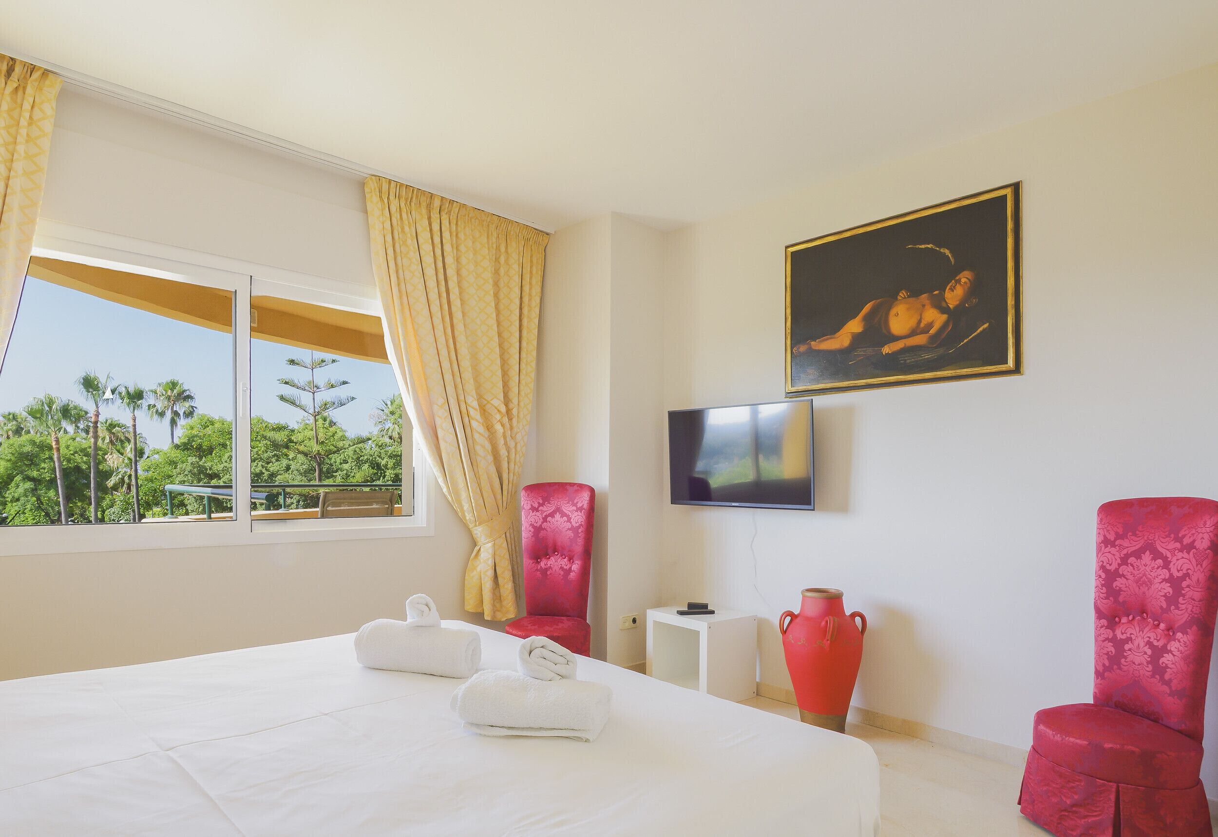 A hotel room with a bed, two red chairs, a TV, and a painting on the wall.
