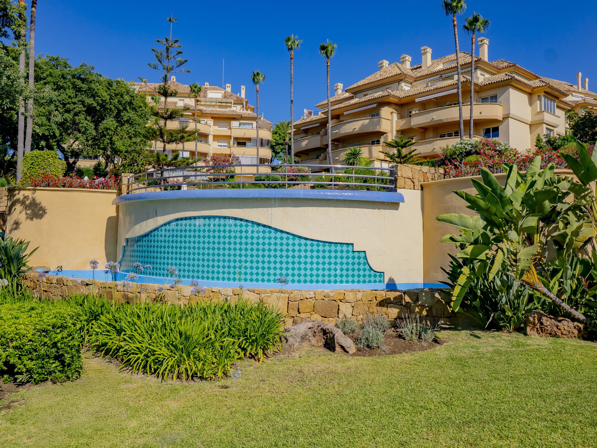 A pool with a blue and white tile pattern, surrounded by a stone wall and lush greenery.