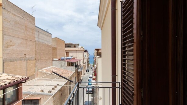 A narrow street with buildings on either side, a visible balcony with a metal railing, and a clear view of the street and buildings in the distance.