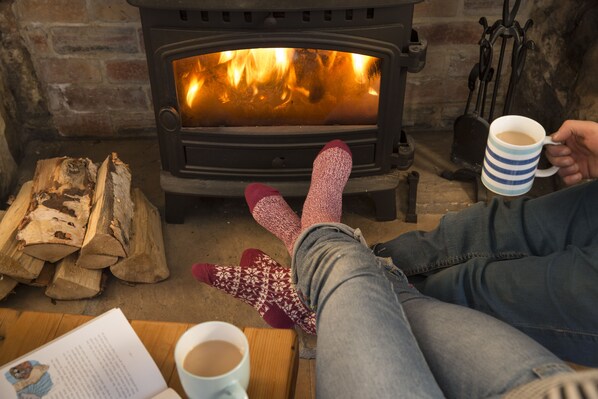 A cozy room with a wood-burning stove, logs, and a person relaxing with a cup of coffee. - Quirky, beach inspired cottage on the fabulous Northumberland Coast. (Amble)