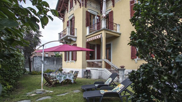 A two-story building with a balcony, outdoor seating area with tables and chairs, and a red umbrella.