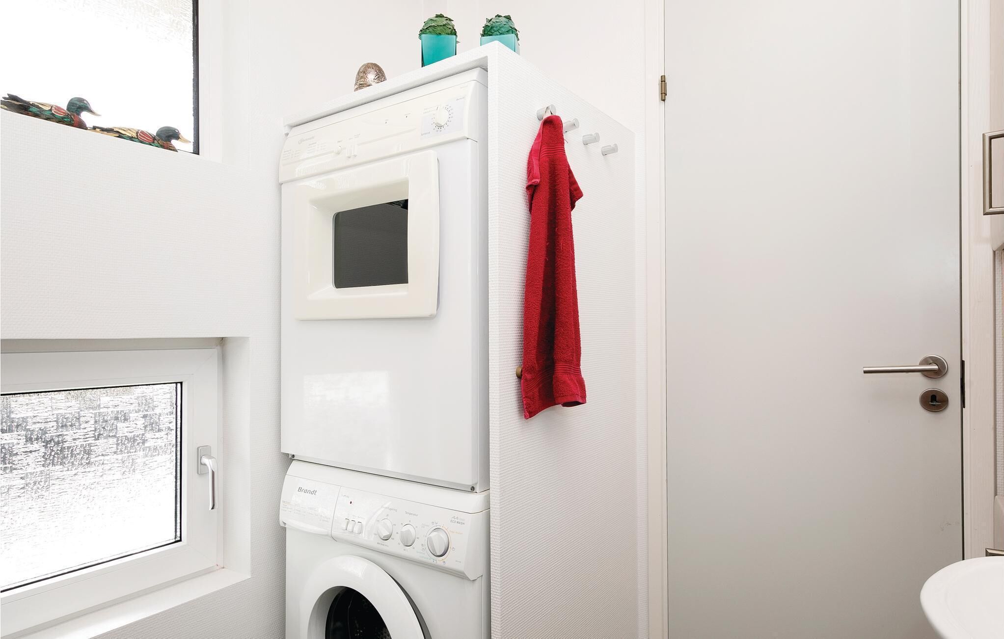 A compact laundry room with a front-loading washing machine, a drying rack with a red towel, and a small window with a view of trees.