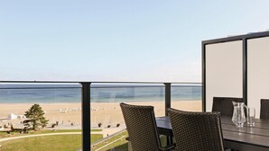 Een balkon met uitzicht op het strand en de oceaan. Er staan rieten stoelen en een tafel met een karaf en glas.