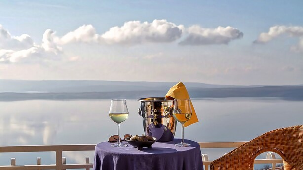 Une table avec un bol de noix, deux verres de vin blanc et un parapluie jaune sur un balcon donnant sur un lac paisible, avec des montagnes à l’horizon.