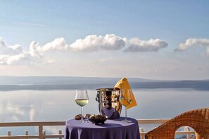 Una mesa con un cuenco de frutos secos, dos copas de vino blanco y un paraguas amarillo en un balcón con vista a un lago tranquilo y montañas a lo lejos.