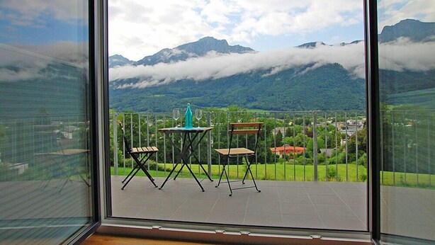 Un balcon avec vue sur les montagnes et les nuages, deux chaises et une table avec une bouteille.