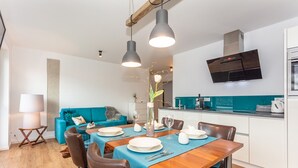 Une cuisine moderne dotée d’un four et d’un micro-ondes encastrés, un coin repas avec une table en bois et une nappe bleue, et un canapé de couleur turquoise en arrière-plan.