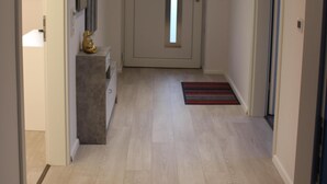 Een hal met een lichtkleurige houten vloer, een witte deur met een verticaal raam en een klein tapijt dat naar een deur aan de rechterkant leidt.