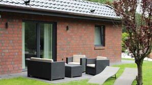 Een modern gebouw met een bakstenen gevel, grote schuifdeuren en terrasmeubilair met zwart riet en beige kussens.