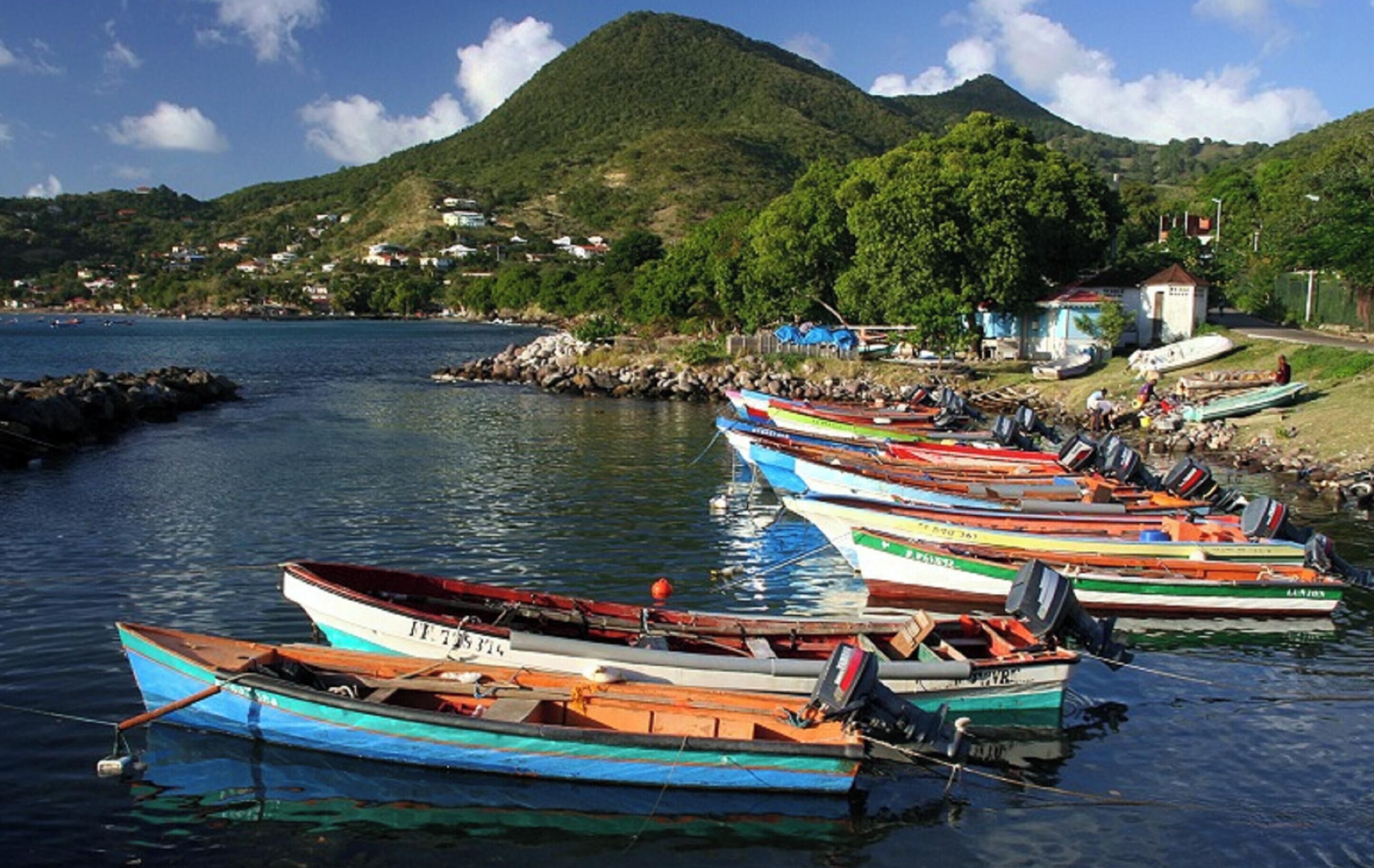 Un port où sont amarrés plusieurs petits bateaux, avec un paysage montagneux en arrière-plan et quelques bâtiments visibles au loin.