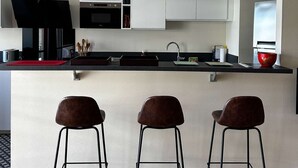 A modern kitchen with a central island, white cabinets, and three bar stools. - Arcachon - Élégant T3 de standing à proximité du centre-ville et plage (ARCACHON)