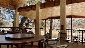 A covered outdoor dining area with a wooden table and chairs, stone pillars, and a view of trees.