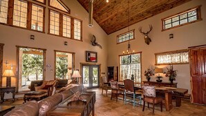 A spacious living room with leather sofas, a wooden coffee table, and a dining area with chairs and a table. Large windows allow natural light, and a Texas flag is displayed on the wall.