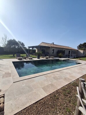 A rectangular swimming pool with a pergola and lounge chairs, surrounded by a patio area with stone tiles.