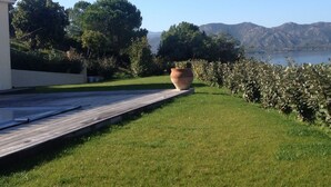 Uma área gramada com um deck de madeira, um grande vaso de terracota e vista para montanhas e um lago.